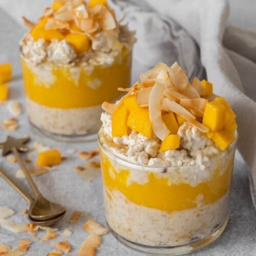 Two glass dishes of creamy bircher with a layer of fresh mango puree, topped with fresh mango chunks, toasted coconut and maple syrup