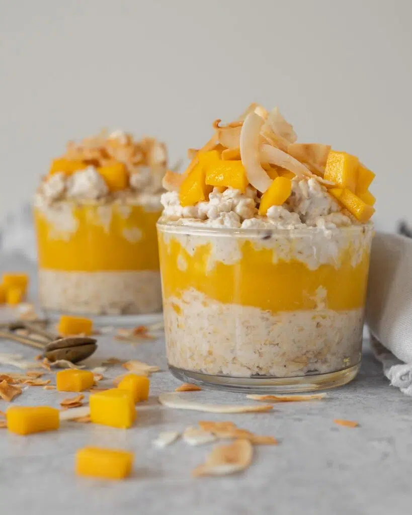 Two glass dishes of creamy bircher with a layer of fresh mango puree, topped with fresh mango chunks, toasted coconut and maple syrup
