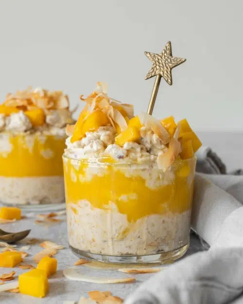 Two glass dishes of mango coconut overnight oats with a layer of fresh mango puree, topped with fresh mango chunks, toasted coconut and maple syrup