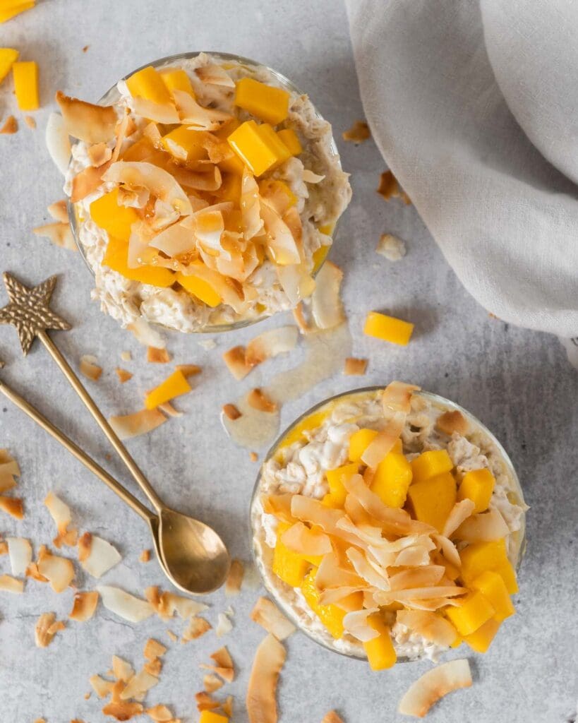 Two glass dishes of creamy bircher with a layer of fresh mango puree, topped with fresh mango chunks, toasted coconut and maple syrup