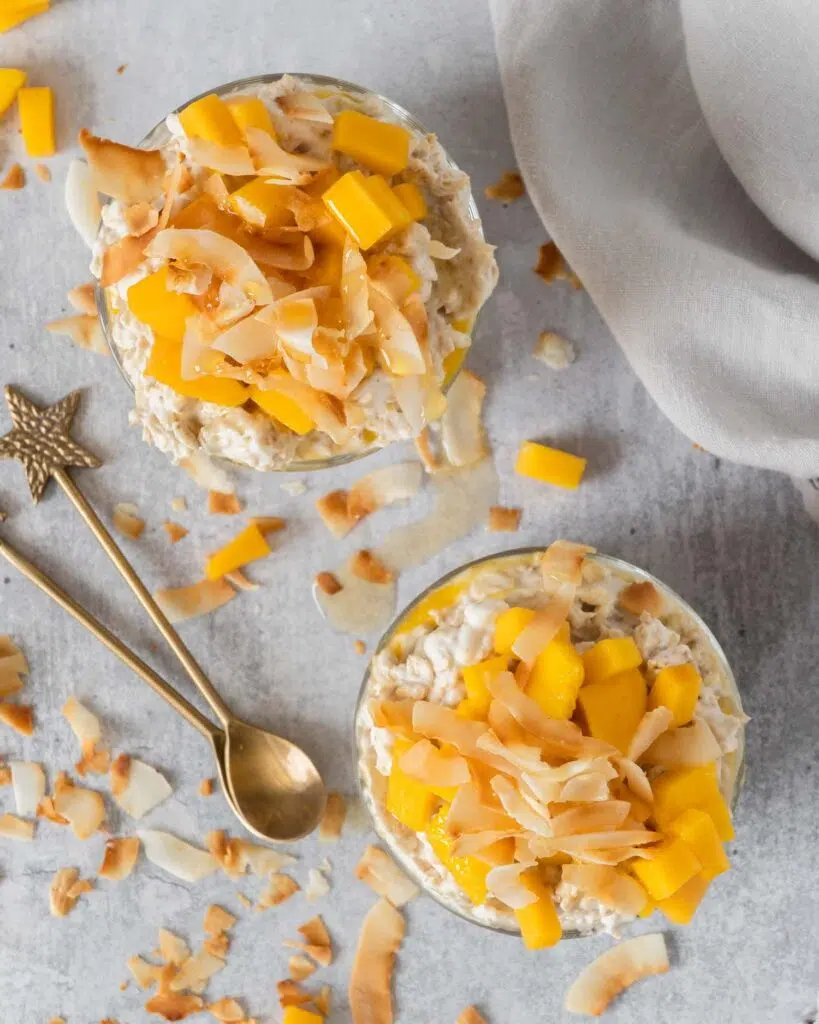 Two glass dishes of creamy bircher with a layer of fresh mango puree, topped with fresh mango chunks, toasted coconut and maple syrup