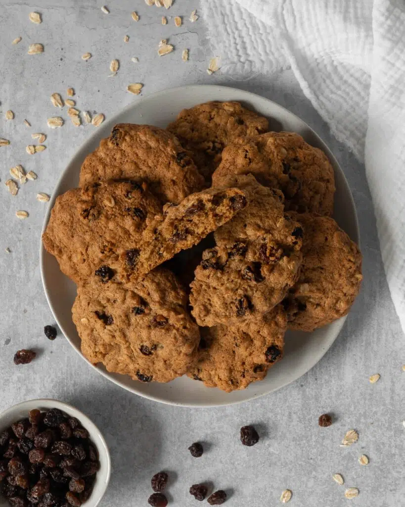 A plate of chunky cinnamon oat raisin cookies with the top cookie broken open to display the soft, squishy interior studded with juicy raisins