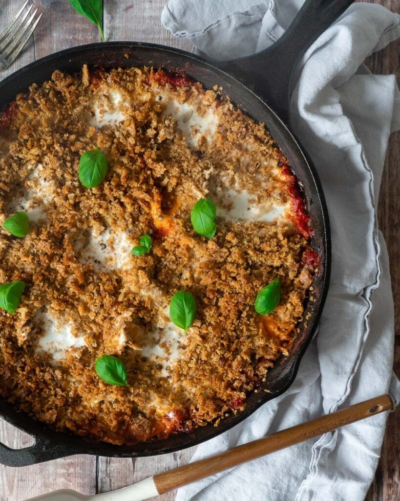 The top of a pan of vegetable lasagne showing a creamy topping with crispy breadcrumbs and fresh basil leaves