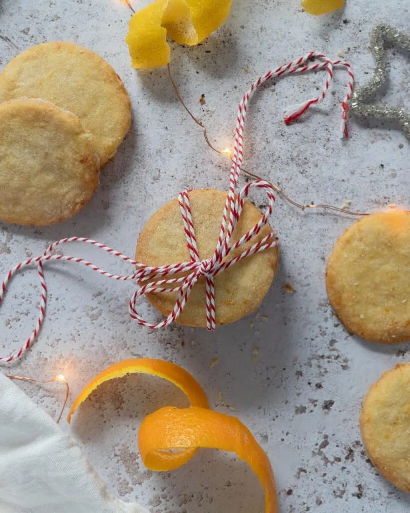 Orange and lemon St Clements shortbread biscuits tied with a red and white striped string and surrounded by fresh orange and lemon peel and fairy lights