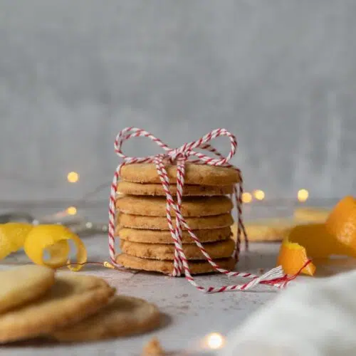 Orange and lemon St Clements shortbread biscuits tied with a red and white striped string and surrounded by fresh orange and lemon peel and fairy lights