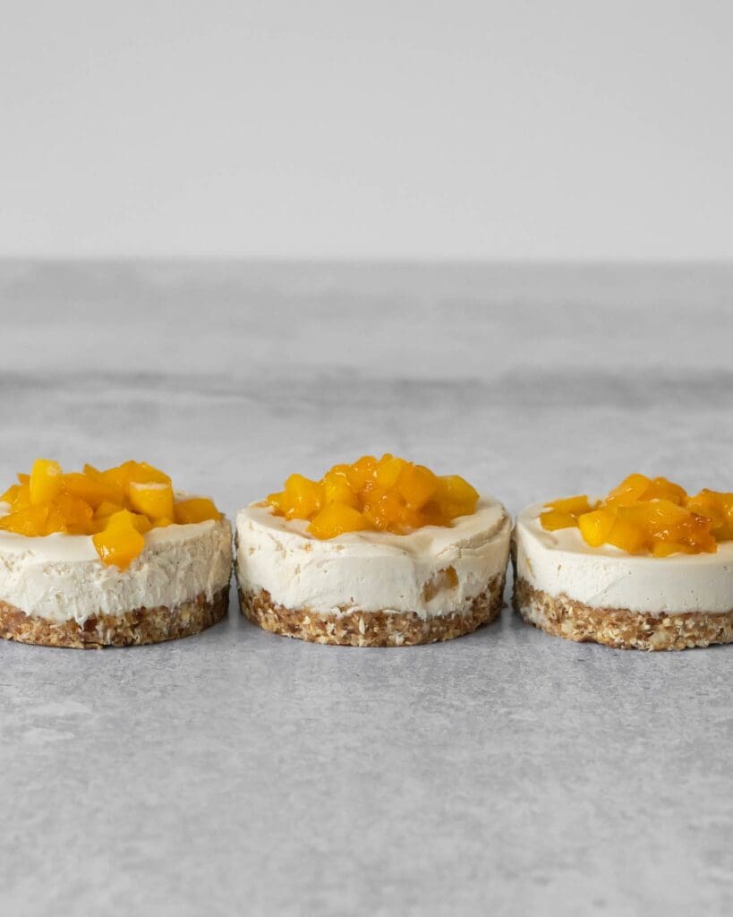 Three individual mini vegan cheesecakes in a row with sticky mango pieces on top
