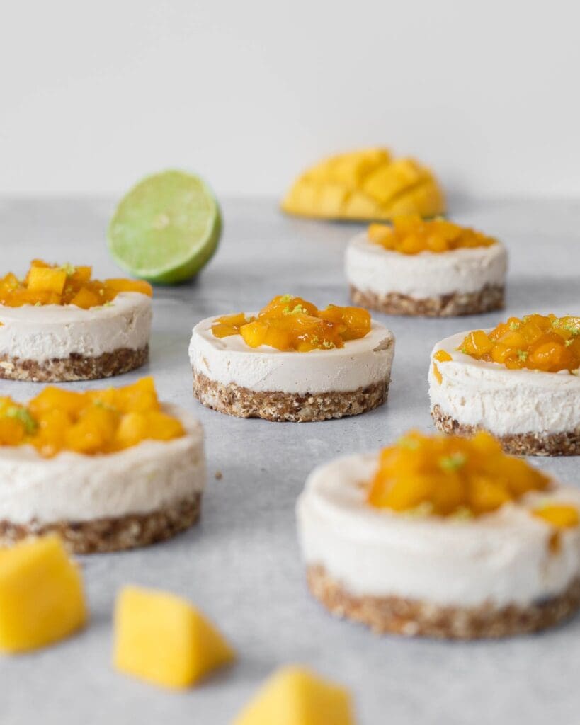 Individual vegan cheesecakes with sticky mango pieces on top sat on a table top with fresh lime and mango scattered around