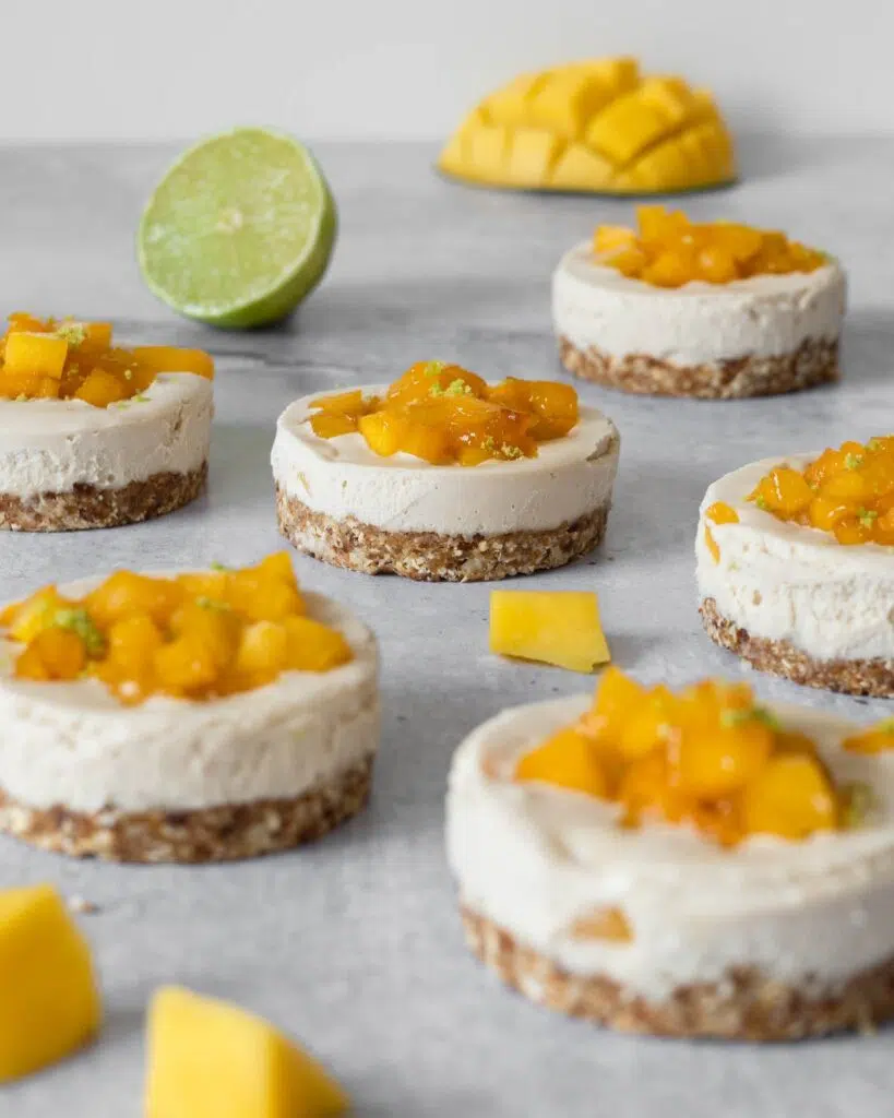 Individual vegan cheesecakes with sticky mango pieces on top sat on a table top with fresh lime and mango scattered around