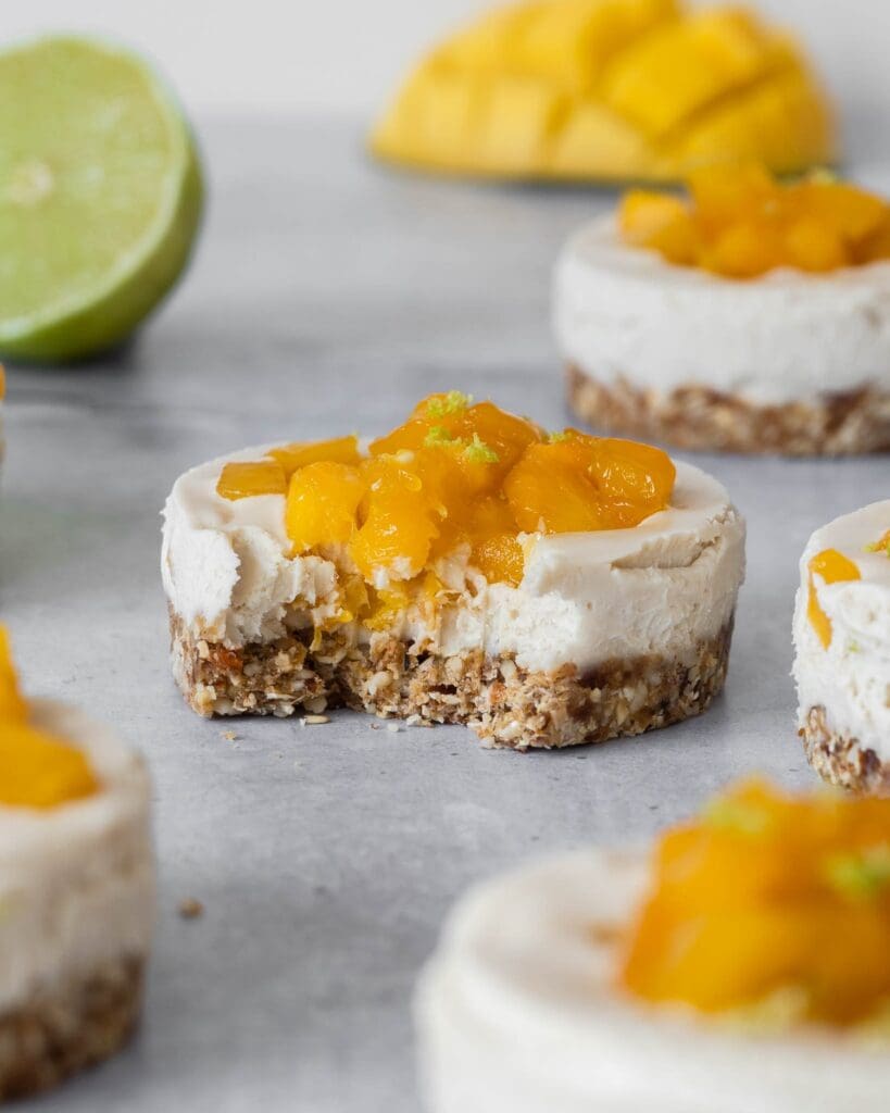 Individual vegan cheesecakes with sticky mango pieces on top sat on a table top with fresh lime and mango scattered around