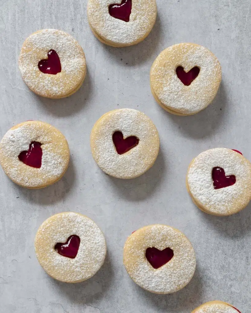 Valentine's Jammie Biscuits on a table top dusted with icing sugar, with heart shaped cut outs displaying the raspberry jam in the centre.