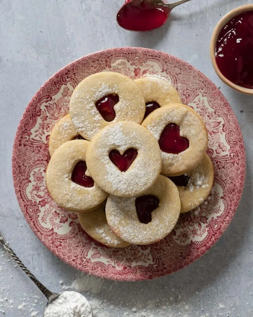 A vintage pink plate of Valentine's Jammie Biscuits dusted with icing sugar, with heart shaped cut outs displaying the raspberry jam in the centre.