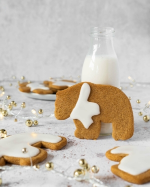 A vegan gingerbread cookie shaped like a bear, wearing a white icing scarf, stood up against a little bottle of oat milk