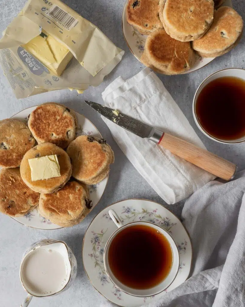 Chunky round vegan welsh cakes studded with juicy sultanas on a plate, on a table with cups of tea, a jug of milk and an open pack of vegan butter