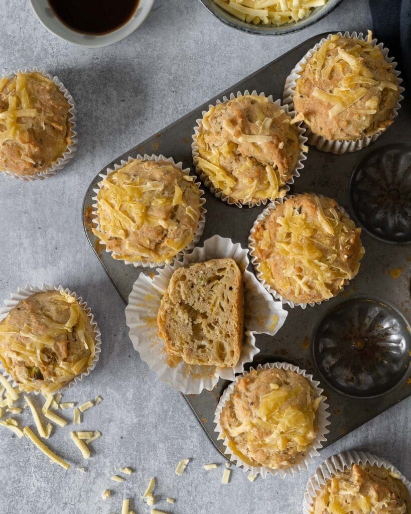 A muffin tray filled with cheesy Welsh Rarebit Muffins, with one muffin cut in half displaying a squishy interior