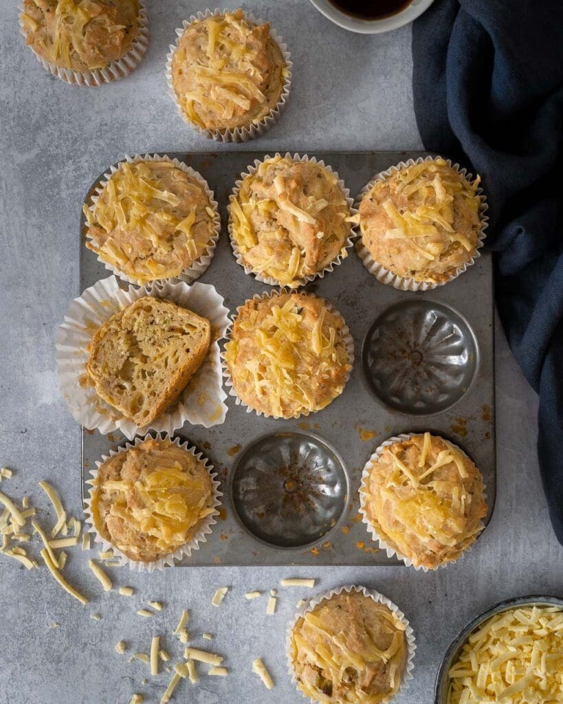 A muffin tray filled with cheesy Welsh Rarebit Muffins, with one muffin cut in half displaying a squishy interior