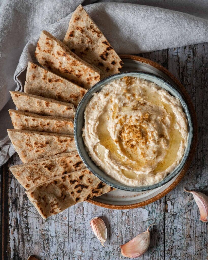 A bowl of creamy white bean hummus whipped to perfection with a drizzle of olive oil on top, a sprinkling of cumin and toasted flat bread strips to dip