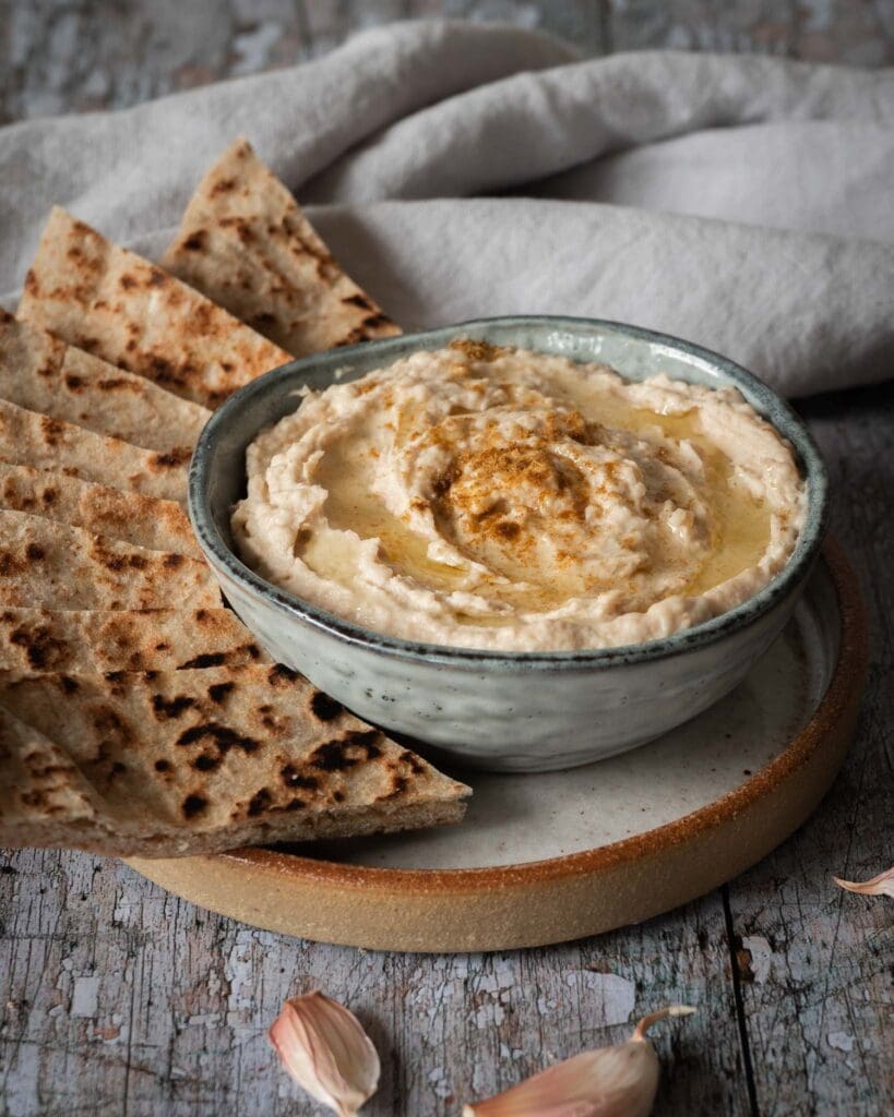A bowl of creamy white bean hummus whipped to perfection with a drizzle of olive oil on top, a sprinkling of cumin and toasted flat bread strips to dip