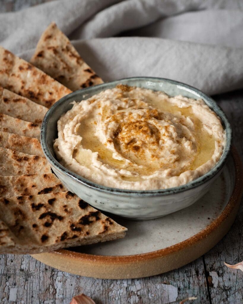 A bowl of creamy white bean hummus whipped to perfection with a drizzle of olive oil on top, a sprinkling of cumin and toasted flat bread strips to dip