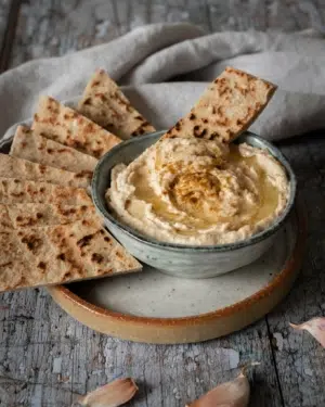 A bowl of creamy white bean hummus whipped to perfection with a drizzle of olive oil on top, a sprinkling of cumin and toasted flat bread strips to dip
