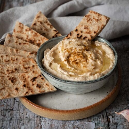 A bowl of creamy white bean hummus whipped to perfection with a drizzle of olive oil on top, a sprinkling of cumin and toasted flat bread strips to dip