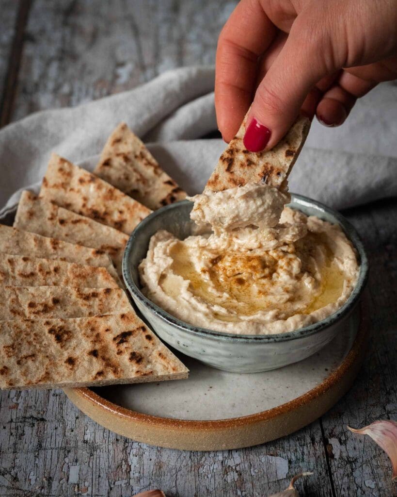 A bowl of creamy white bean hummus whipped to perfection with a drizzle of olive oil on top, a sprinkling of cumin and toasted flat bread strips to dip