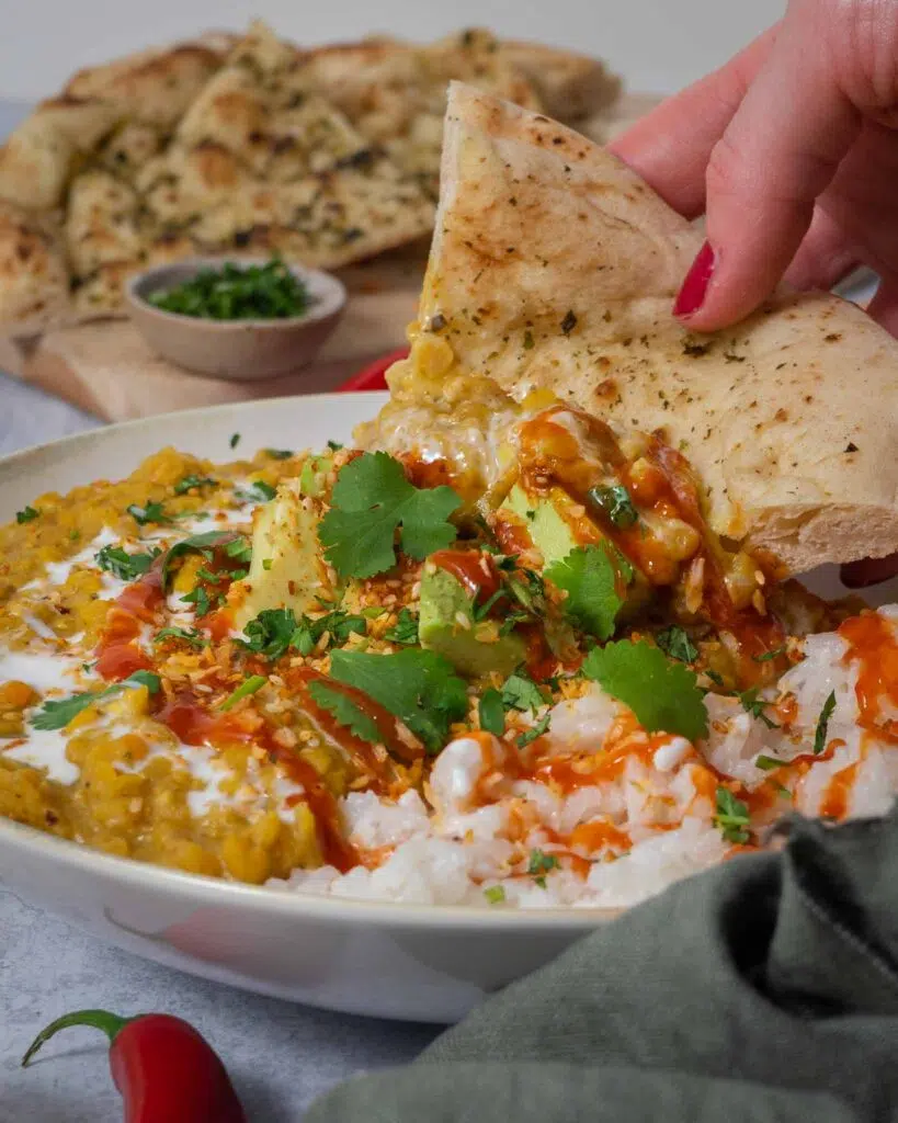 A slice of naan bread being dipped into a hearty bowl of creamy vegan red lentil curry topped with avocado, coriander and chilli sauce.