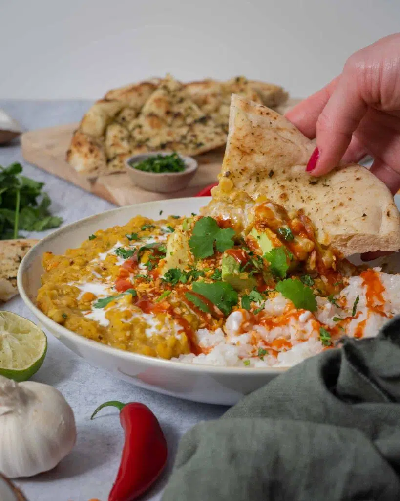 A slice of naan bread being dipped into a hearty bowl of vegan red lentil dahl topped with creamy avocado, coriander and chilli sauce.