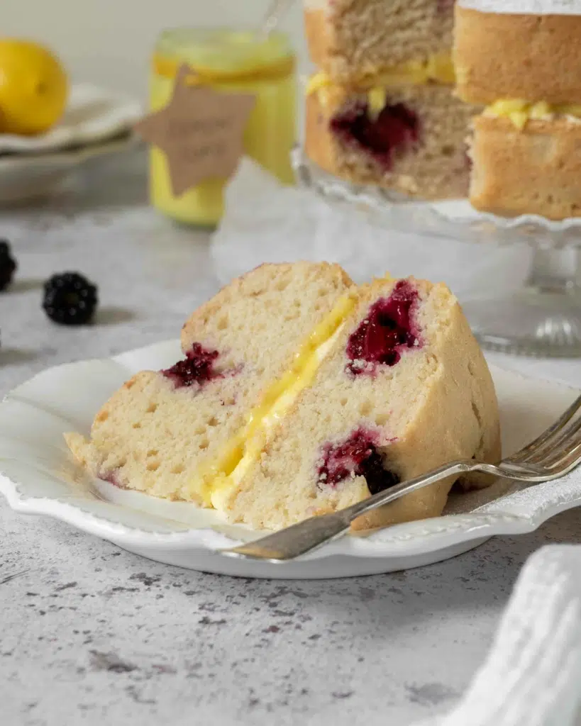 A slice of lemon sponge cake, dotted with fresh blackberries and filled with lemon curd and lemon buttercream.