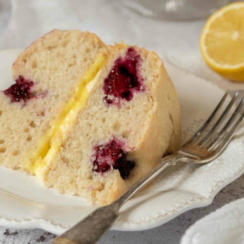 A slice of lemon sponge cake, dotted with fresh blackberries and filled with lemon curd and lemon buttercream.