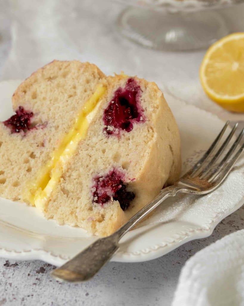 A slice of lemon sponge cake, dotted with fresh blackberries and filled with lemon curd and lemon buttercream.