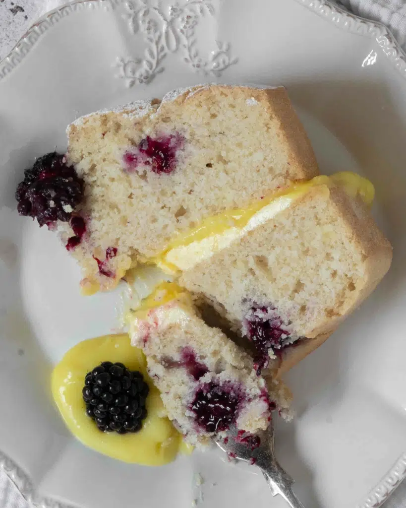 A slice of lemon sponge cake, dotted with fresh blackberries and filled with lemon curd and lemon buttercream.