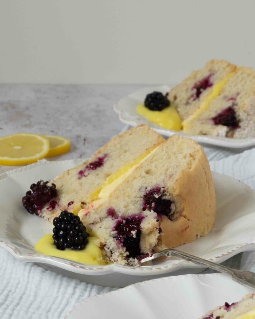 A slice of lemon sponge cake, dotted with fresh blackberries and filled with lemon curd and lemon buttercream.