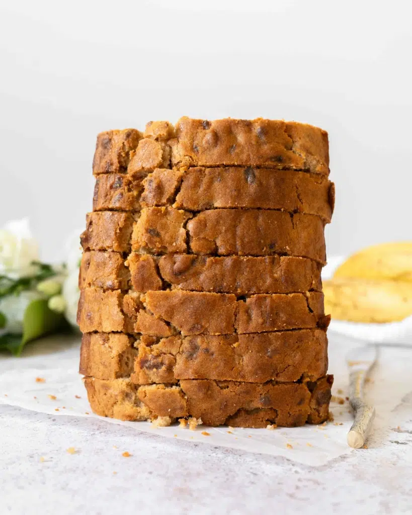 A stack of banana bread slices, golden and enticing. With ripe bananas and fresh white flowers out of focus in the background.