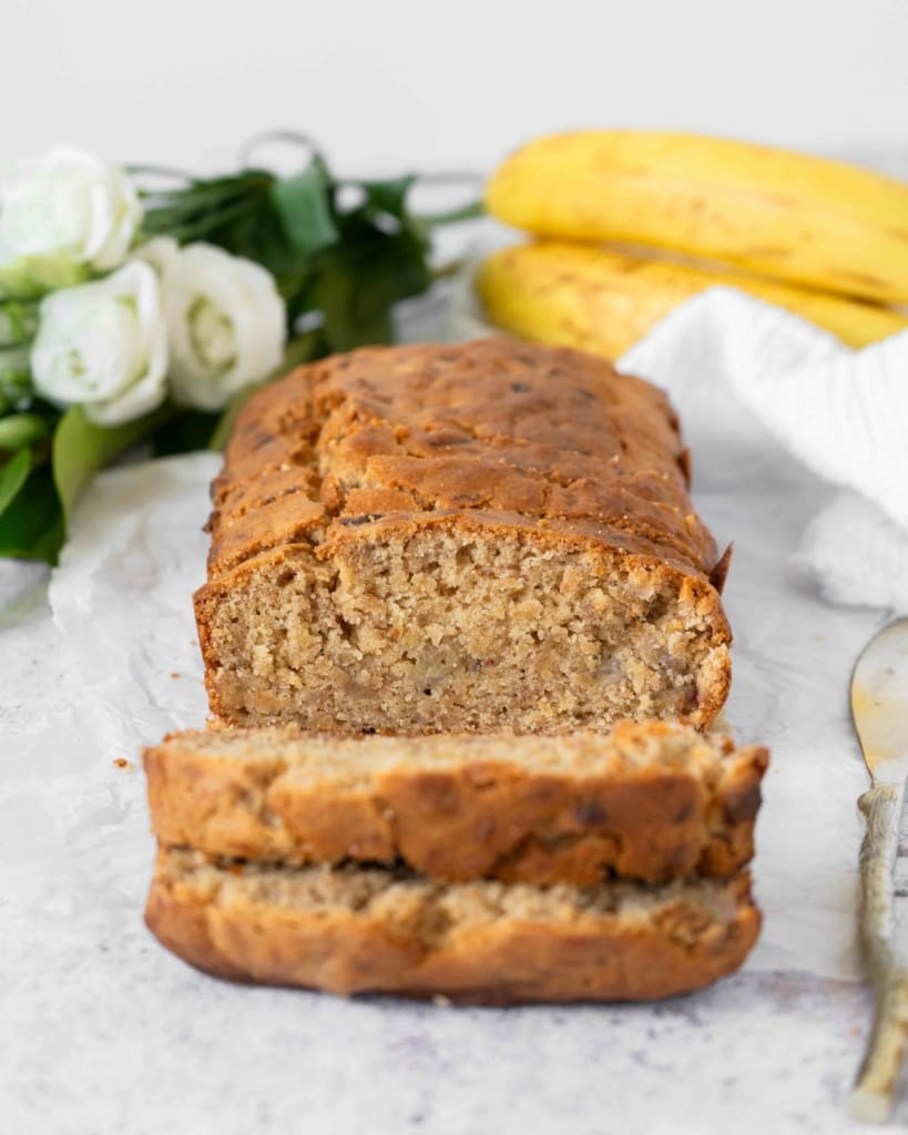 A loaf of banana bread laying on a table, with two slices cut, revealing the soft, tender crumb of the inside of the cake. In the background are white flowers and ripe bananas laying on the table, out of focus.