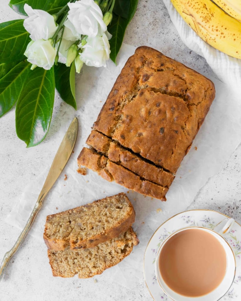 An overhead photograph of a loaf of banana bread on a table top. Two slices of banana bread have been cut and lay next to a cup of tea, fresh flowers and ripe bananas.