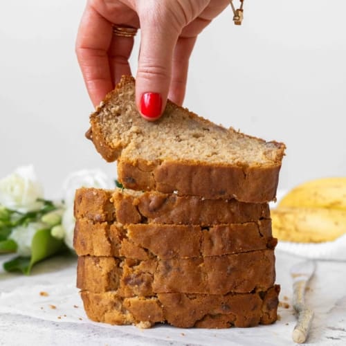 A stack of 5 ingredient vegan banana bread slices, golden and enticing. With ripe bananas and fresh white flowers out of focus in the background. The top slice of banana bread is being picked up to display the tender crumb.