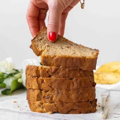 A stack of banana bread slices, golden and enticing. With ripe bananas and fresh white flowers out of focus in the background. The top slice of banana bread is being picked up to display the tender crumb.