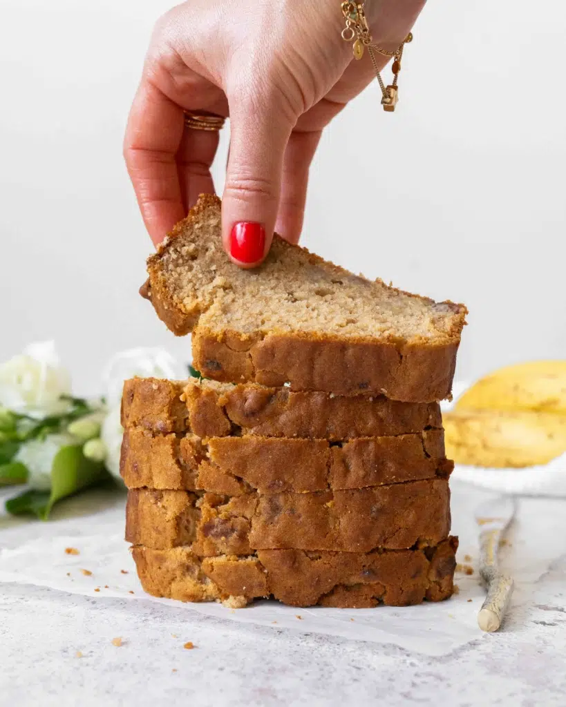 A stack of banana bread slices, golden and enticing. With ripe bananas and fresh white flowers out of focus in the background. The top slice of banana bread is being picked up to display the tender crumb.