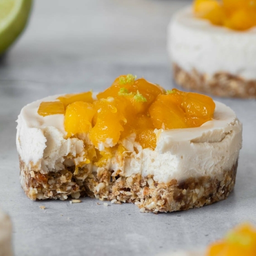 Individual vegan cheesecakes with sticky mango pieces on top sat on a table top with fresh lime and mango scattered around