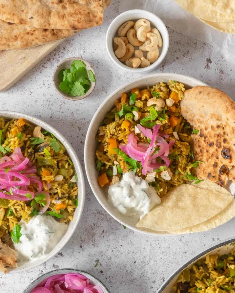 An overhead view of two bowls of vegan biryani curry, topped with mint and cucumber raita dip, pickled red onions, fresh coriander and toasted cashew nuts.