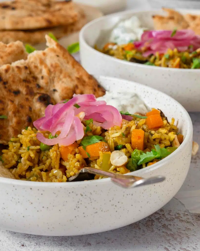 A bowl of vegan spiced rice biryani, topped with mint and cucumber raita dip, pickled red onions, fresh coriander, toasted cashew nuts, poppadoms and naan bread. The biryani is full of fresh and colourful vegetables including diced carrot and green pepper.