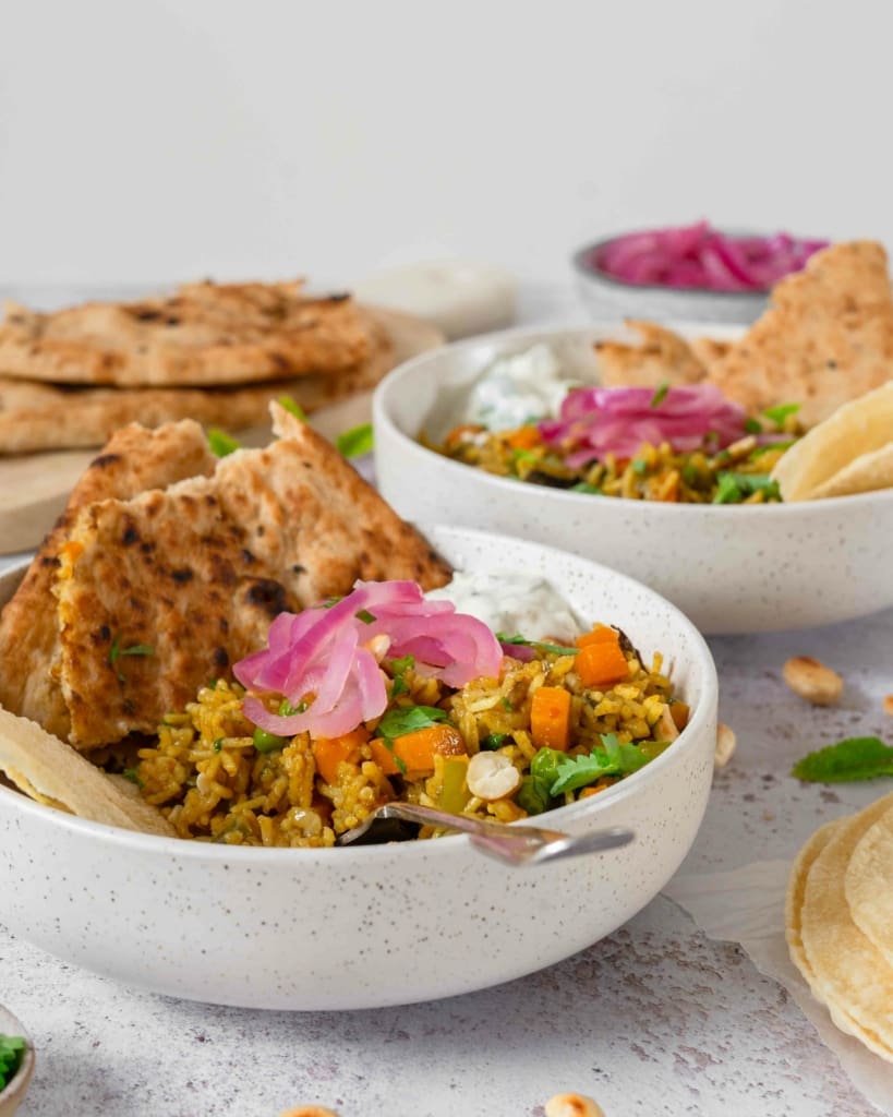 Two bowls of vegan biryani curry, topped with mint and cucumber raita dip, pickled red onions, fresh coriander, toasted cashew nuts, poppadoms and naan bread.