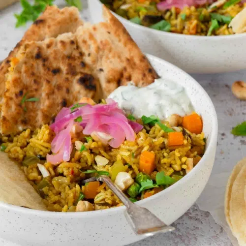 A close up photograph of a bowl of vegetable biryani, topped with mint and cucumber raita dip, pickled red onions, fresh coriander, toasted cashew nuts, poppadoms and naan bread. The biryani is full of fresh and colourful vegetables including diced carrot and green pepper.