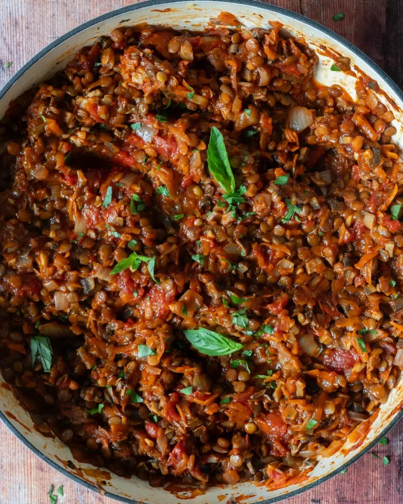 A large pan of rich vegan mushroom and lentil bolognese on the stove top, with fresh basil leaves sprinkled on top.