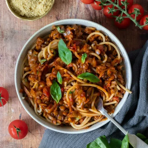 A bowl of rich and flavoursome vegan spaghetti bolognese sat on a table top, surrounded by fresh basil leaves, cherry tomatoes and vegan parmesan.
