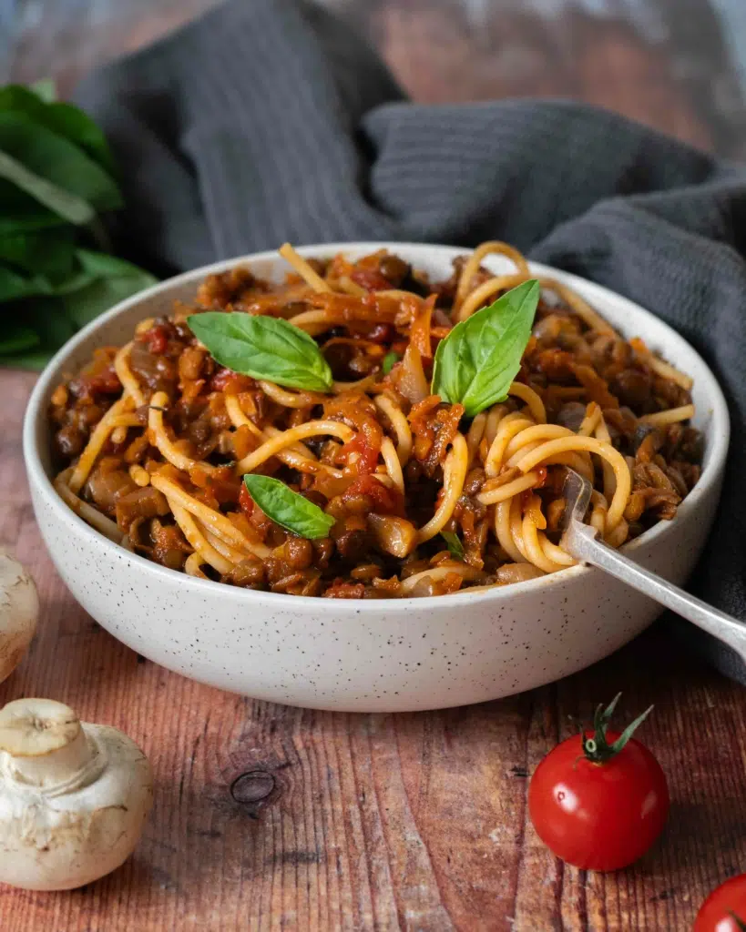 A bowl of rich and flavoursome vegan spaghetti bolognese sat on a table top, topped with fresh basil leaves.