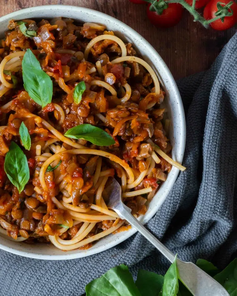 A bowl of rich and flavoursome vegan spaghetti bolognese sat on a table top, topped with fresh green basil leaves.