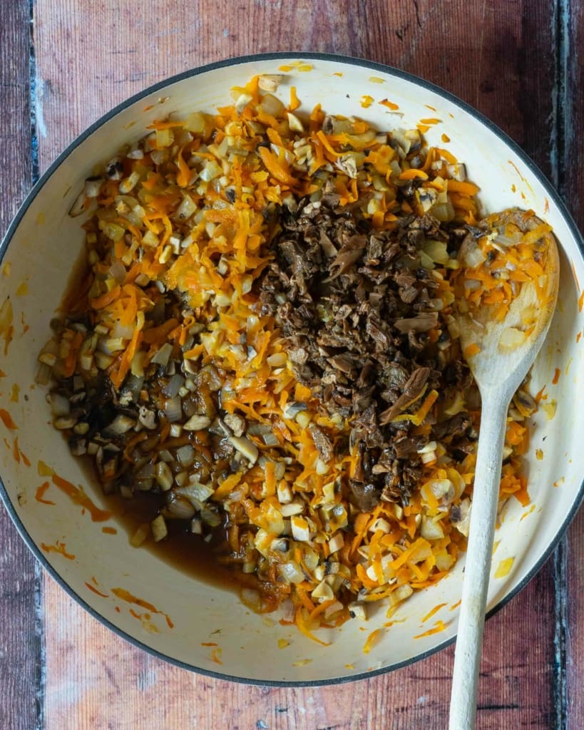 Wild mushrooms being added to a pan of fried carrot, onion, garlic and fresh white mushrooms