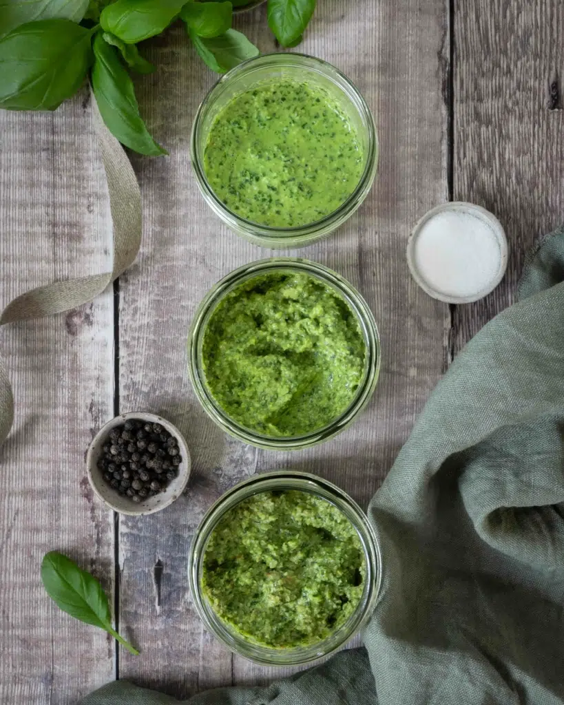 An overhead photo of three bowls of vibrant green vegan pesto. Fresh basil leaves, peppercorns, sea salt and a green napkin surround the bowls.