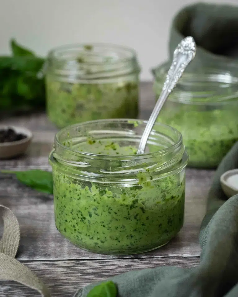 A glass bowl of vibrant green vegan pesto sat on a rustic wooden table top, with fresh basil leaves scattered around. Two bowls of pesto sit in the background - one made without pine nuts, the other is nut free.